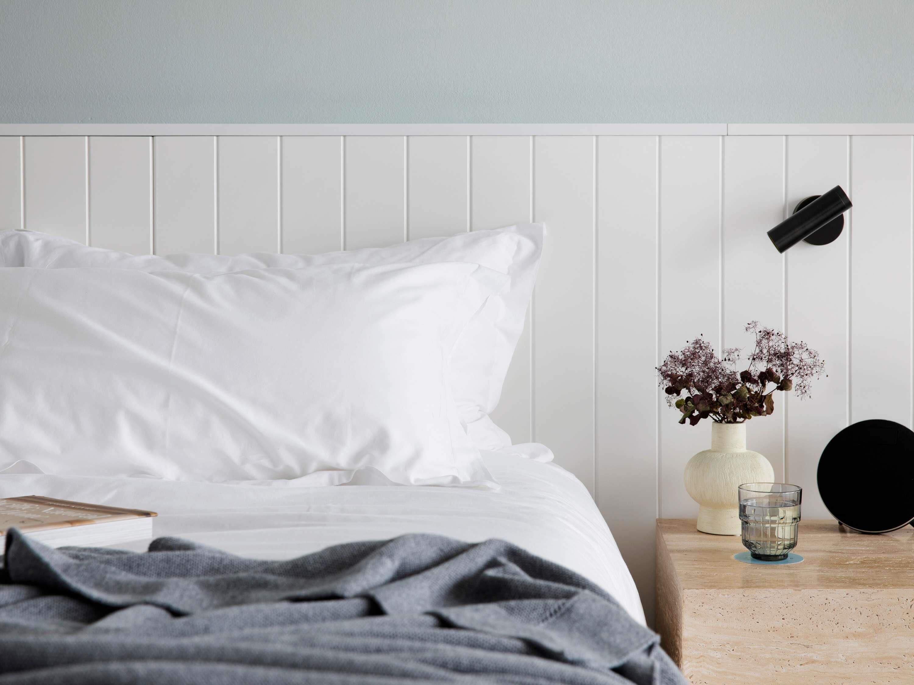 Una cama bien hecha con ropa blanca, un plaid gris, una mesita de noche con un florero, un vaso de agua y un objeto negro en la pared.