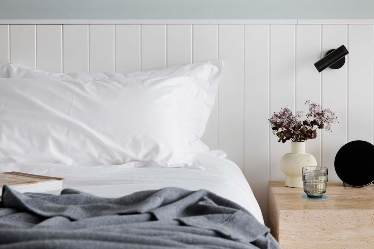 Una cama bien hecha con ropa blanca, un plaid gris, una mesita de noche con un florero, un vaso de agua y un objeto negro en la pared.