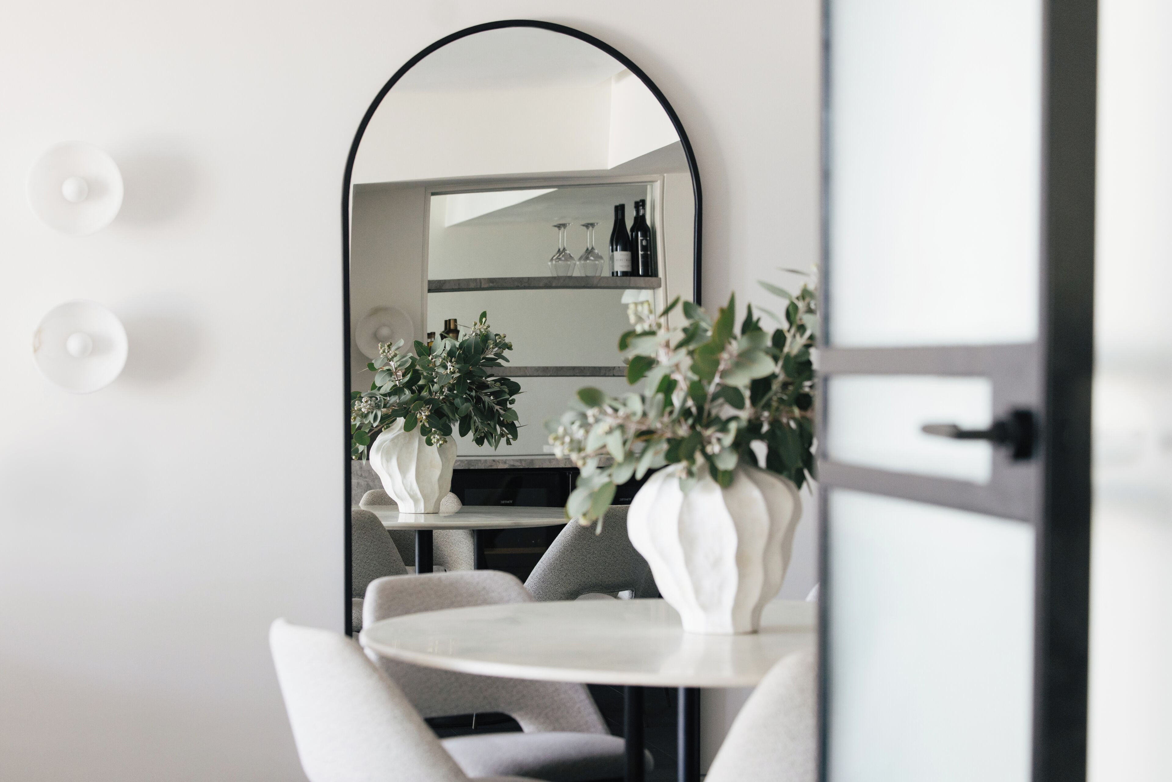 A modern interior with a round table, two potted plants, a mirror, and a shelf with bottles.