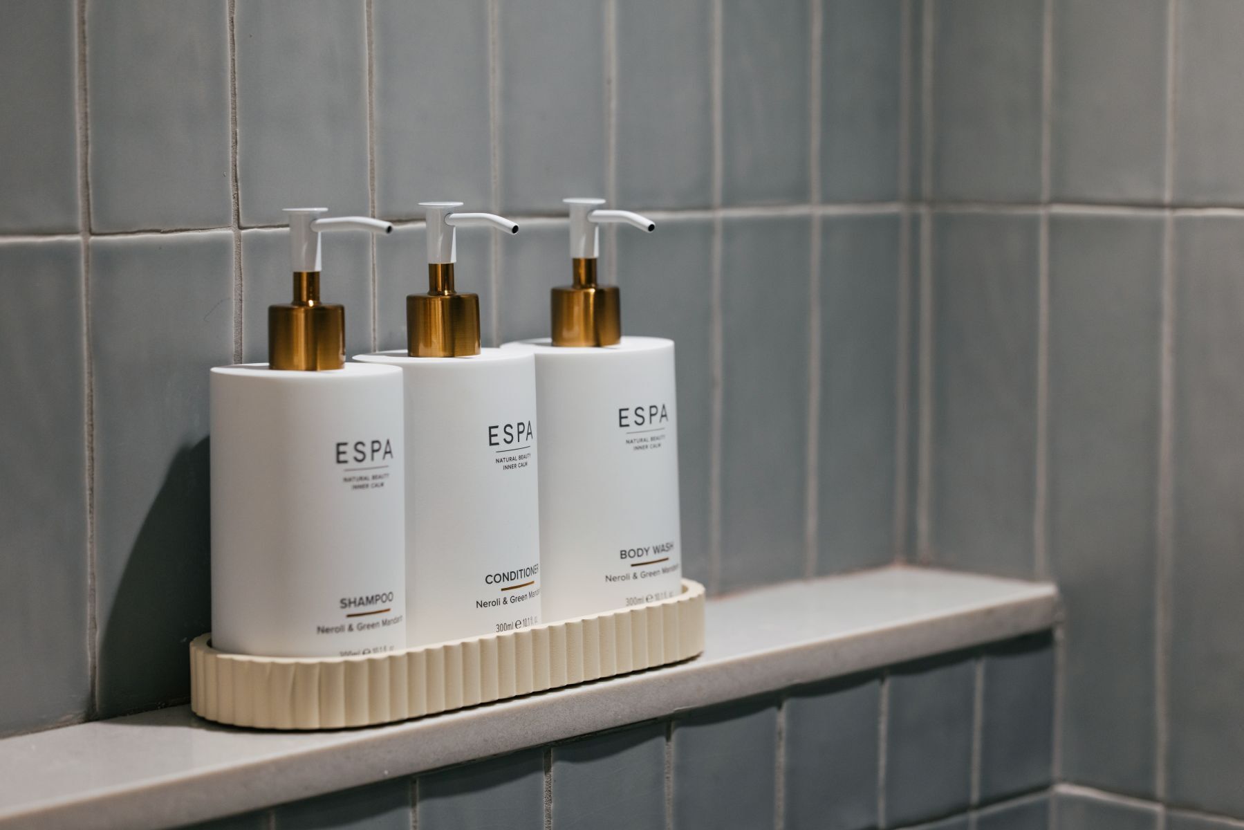 Drie witte flessen met gouden pompen op een witte plank voor een betegelde muur.