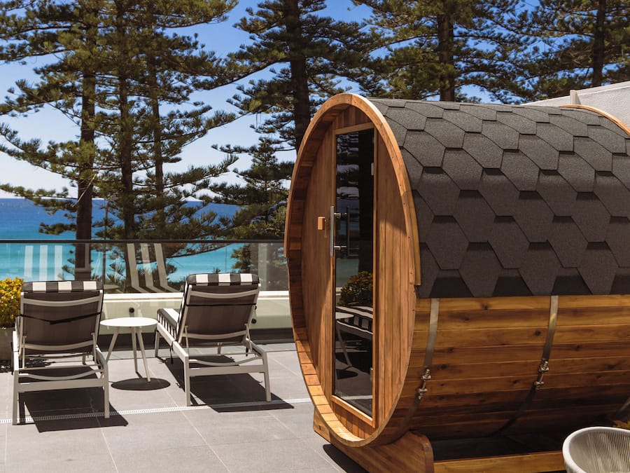 Una estructura de madera con forma de barril, con una puerta, rodeada de asientos exteriores y vistas a árboles y un edificio al fondo.