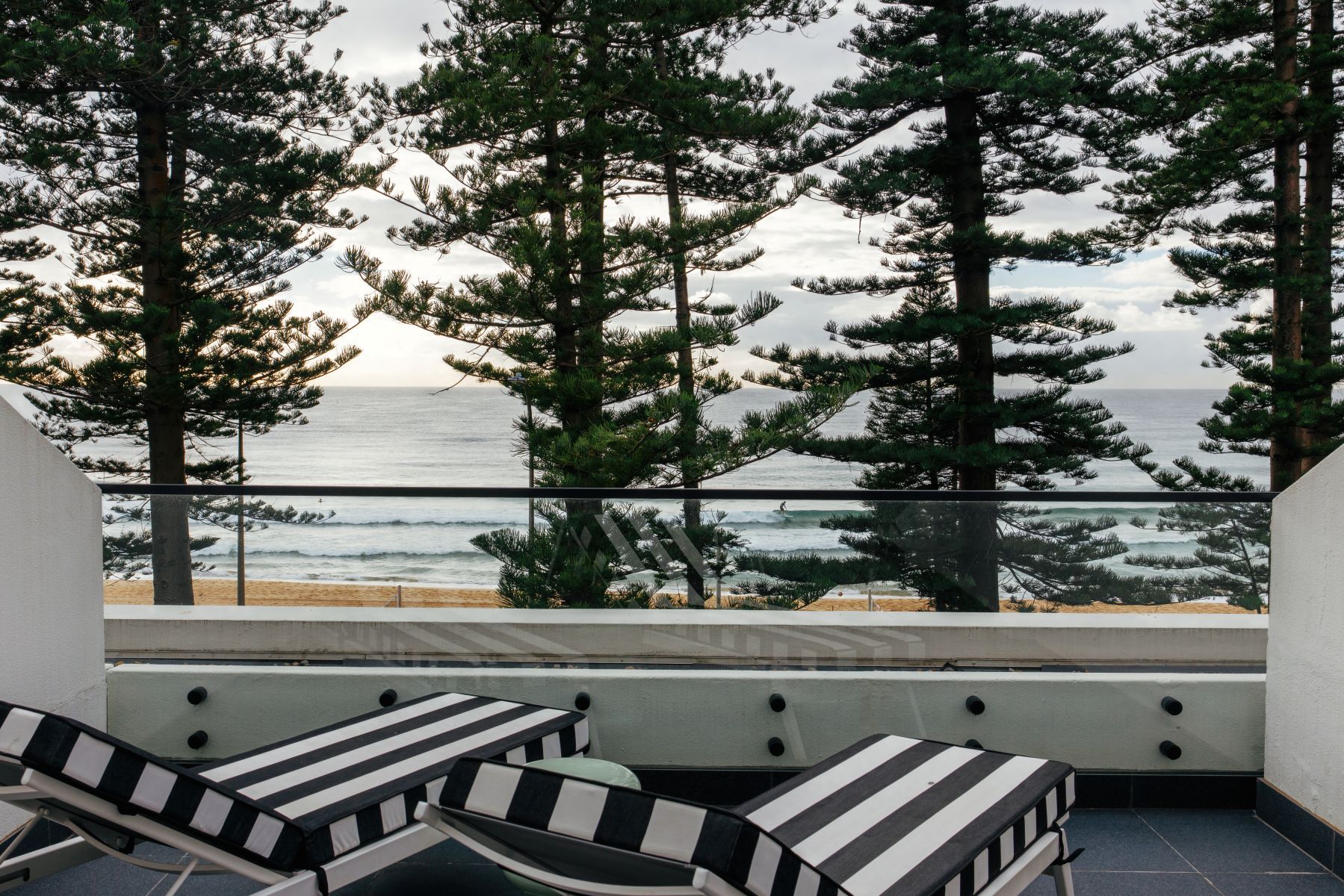 A balcony with striped chairs and a view of the ocean through tall pine trees.