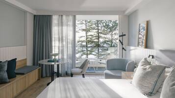 Habitación de hotel con cama, zona de estar con mesa y balcón con vistas a árboles.