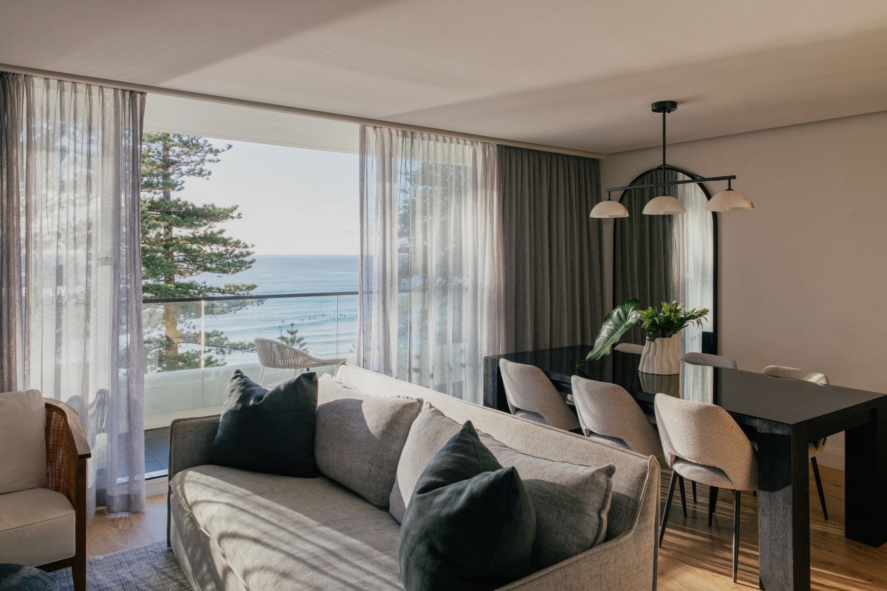 A modern living room with a sofa, dining table, and chairs, overlooking a scenic view of the ocean and trees.