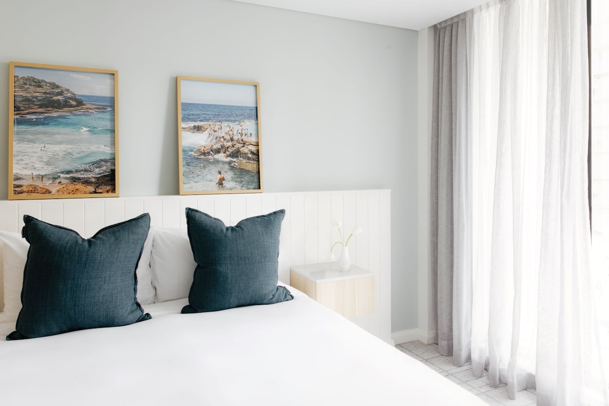 Un dormitorio con cama, dos almohadas, una mesita de noche, un jarrón con flores y dos cuadros en la pared.