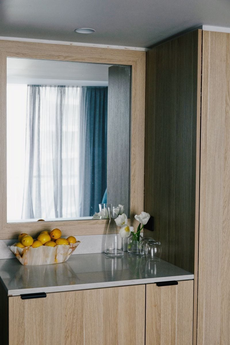 Un comptoir de cuisine avec un bol de citrons, un vase rempli de fleurs blanches et un miroir qui reflète une fenêtre avec des rideaux.