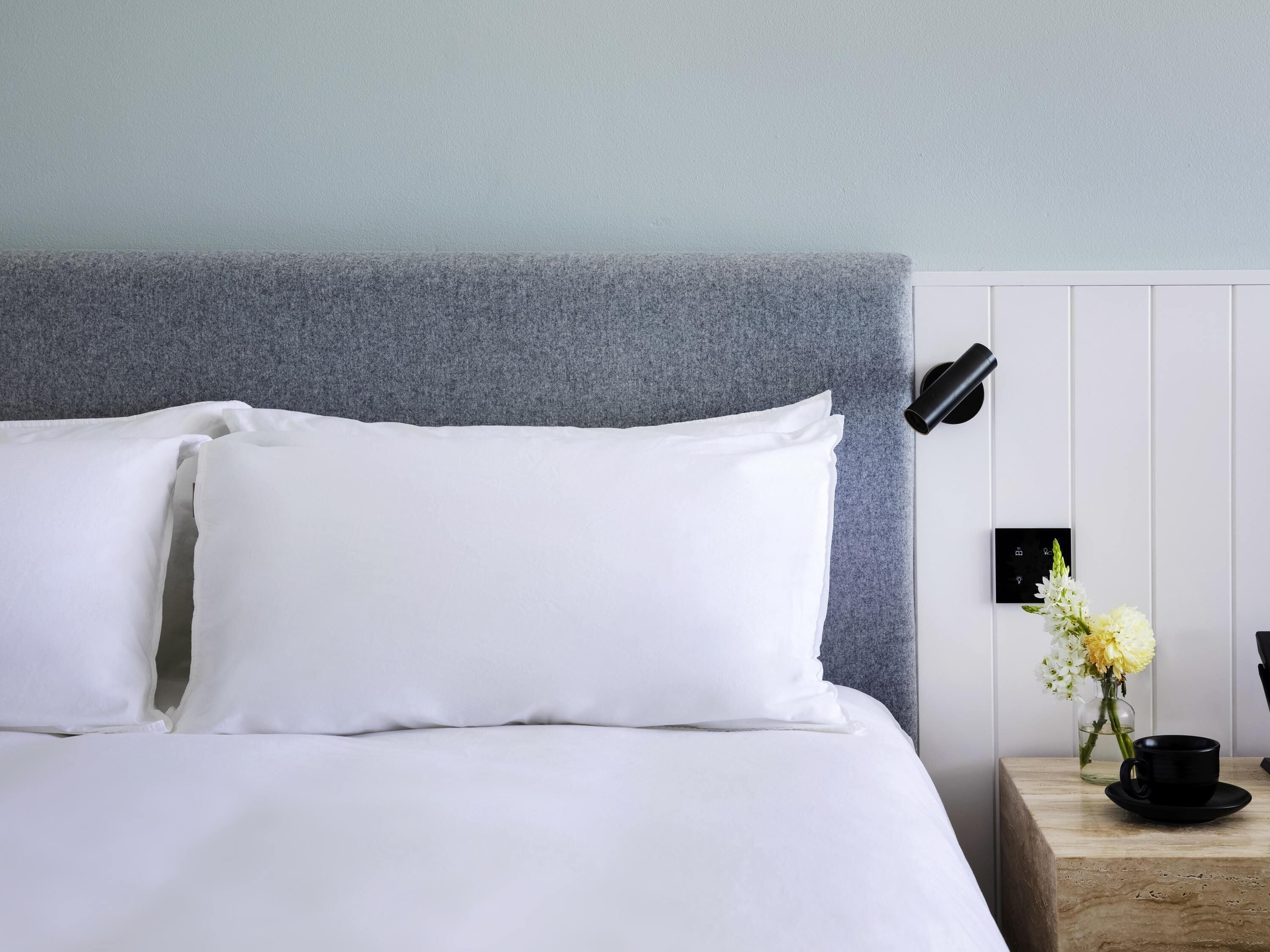 A neatly made bed with white linens, a grey headboard, a bedside table with a vase of flowers, and a black lamp.