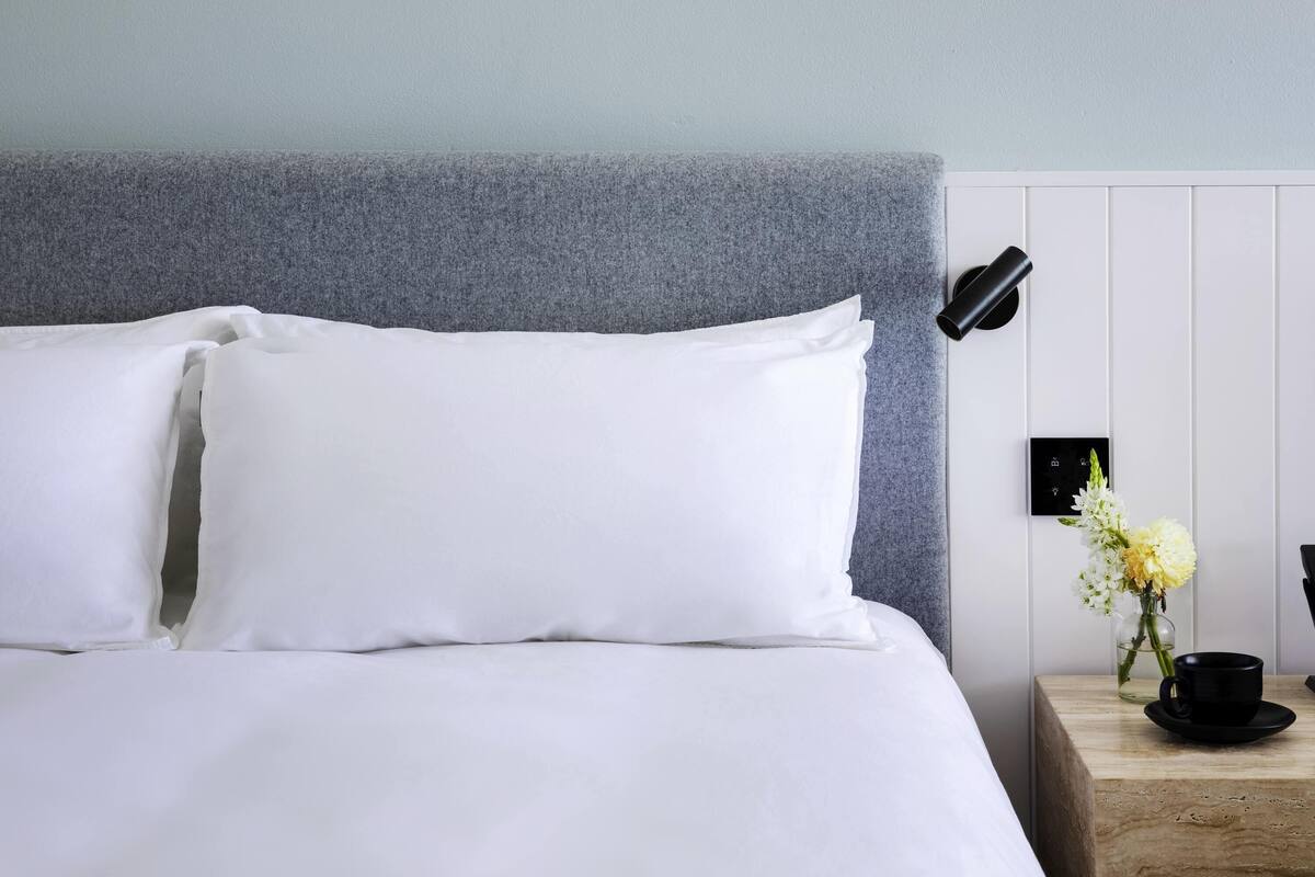 Una cama bien hecha con sábanas blancas, un cabecero gris, una mesita de noche con un florero y una lámpara negra.
