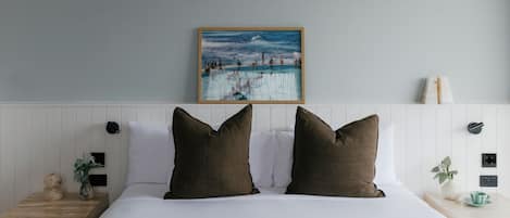 Cama com roupa de cama branca e duas almofadas castanhas, um quadro de uma praia acima e uma parede branca com cabeceira de madeira.