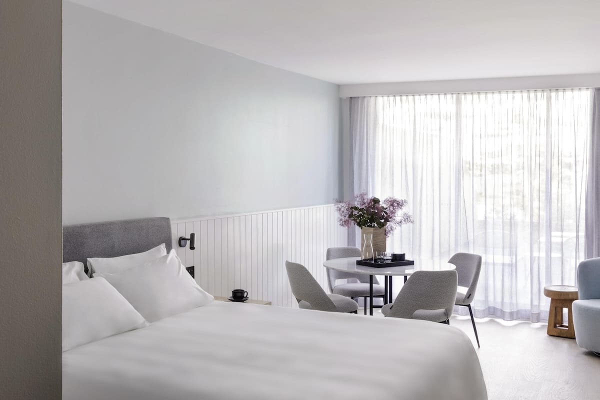 A hotel room with a large bed, a small round table with chairs, a vase of flowers, and a view of the city through sheer curtains.