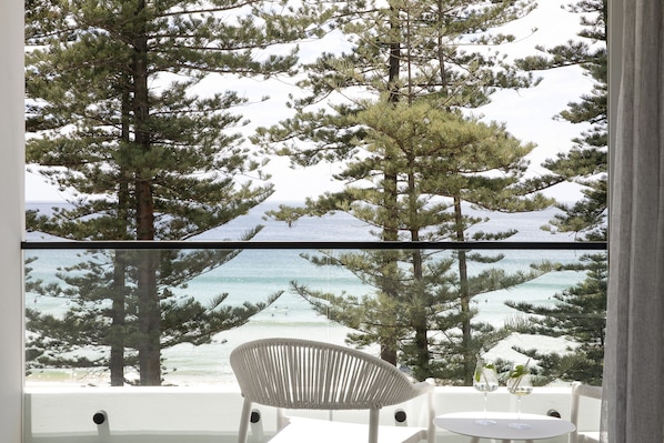 A room with a view of a beach and ocean through large windows, a white chair, and a small table with a glass. - Elysium Manly (Manly)