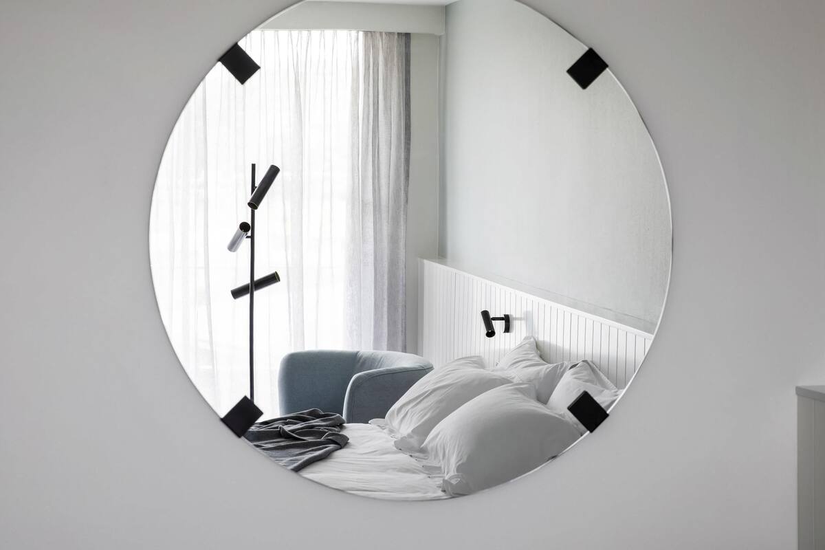 A circular mirror reflecting a bedroom with a bed, pillows, a chair, and a floor lamp.