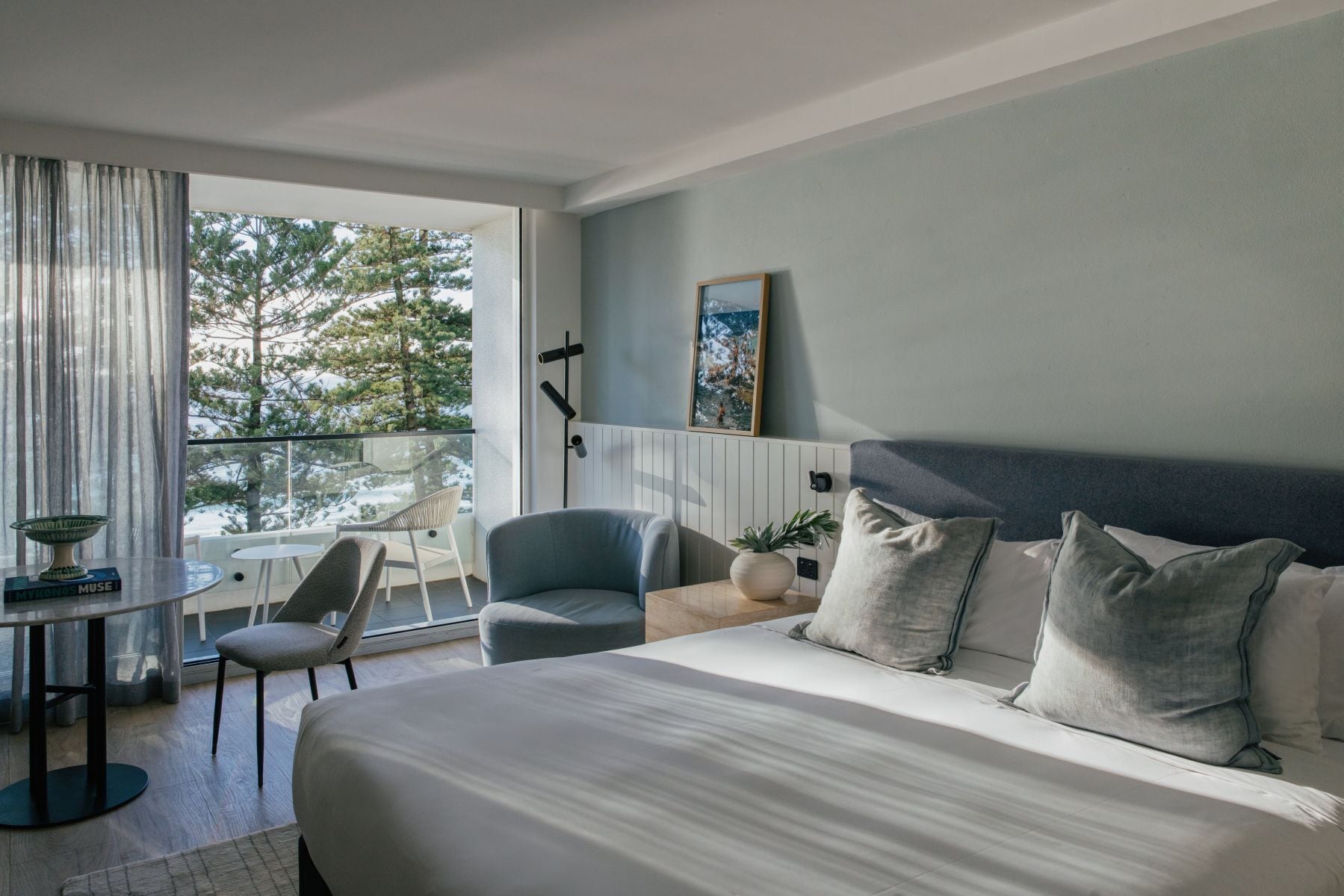 A hotel room with a large bed, a seating area with a table and chairs, a view of trees through the window, and a painting on the wall.
