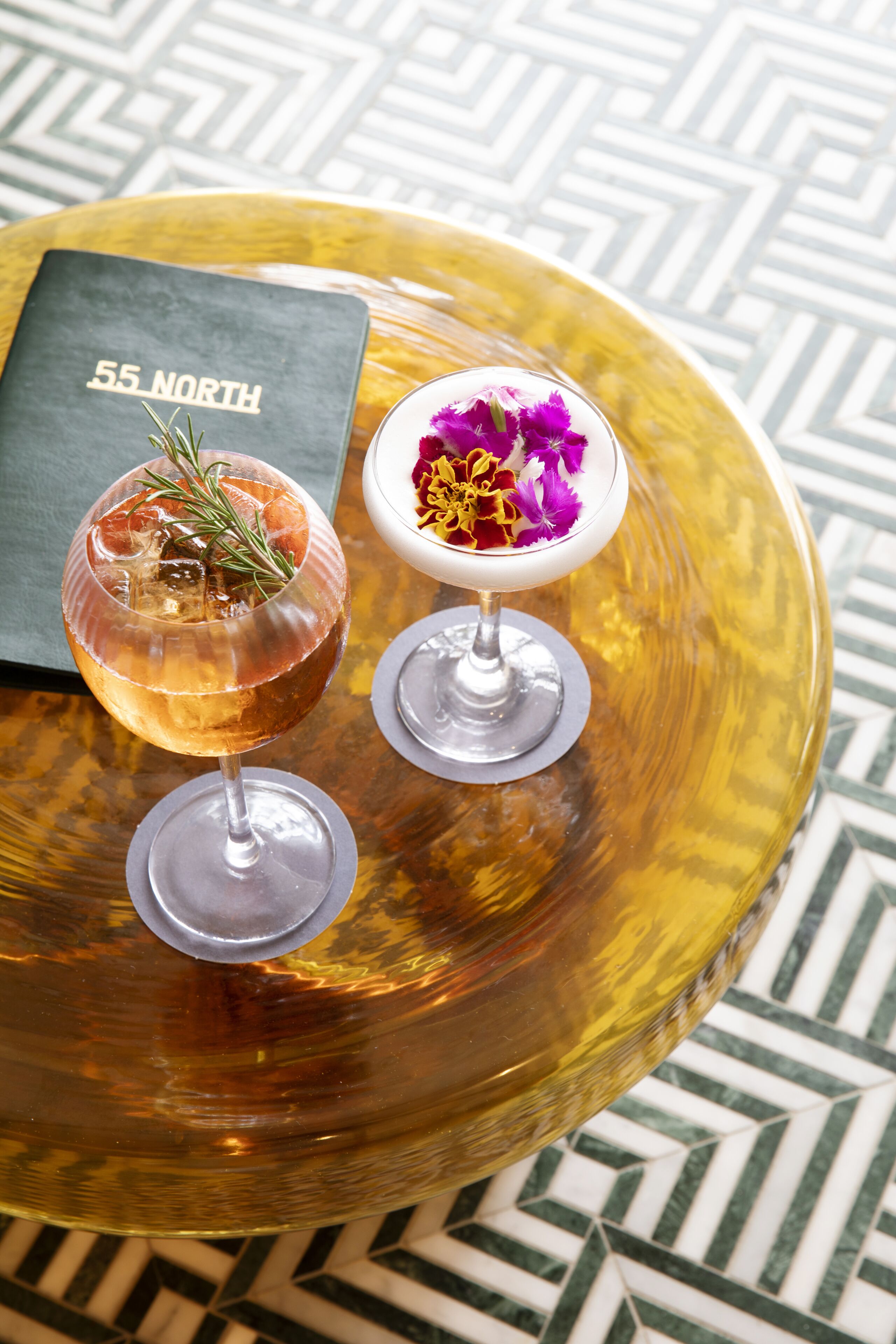 Two cocktails on a round glass table with a patterned floor.