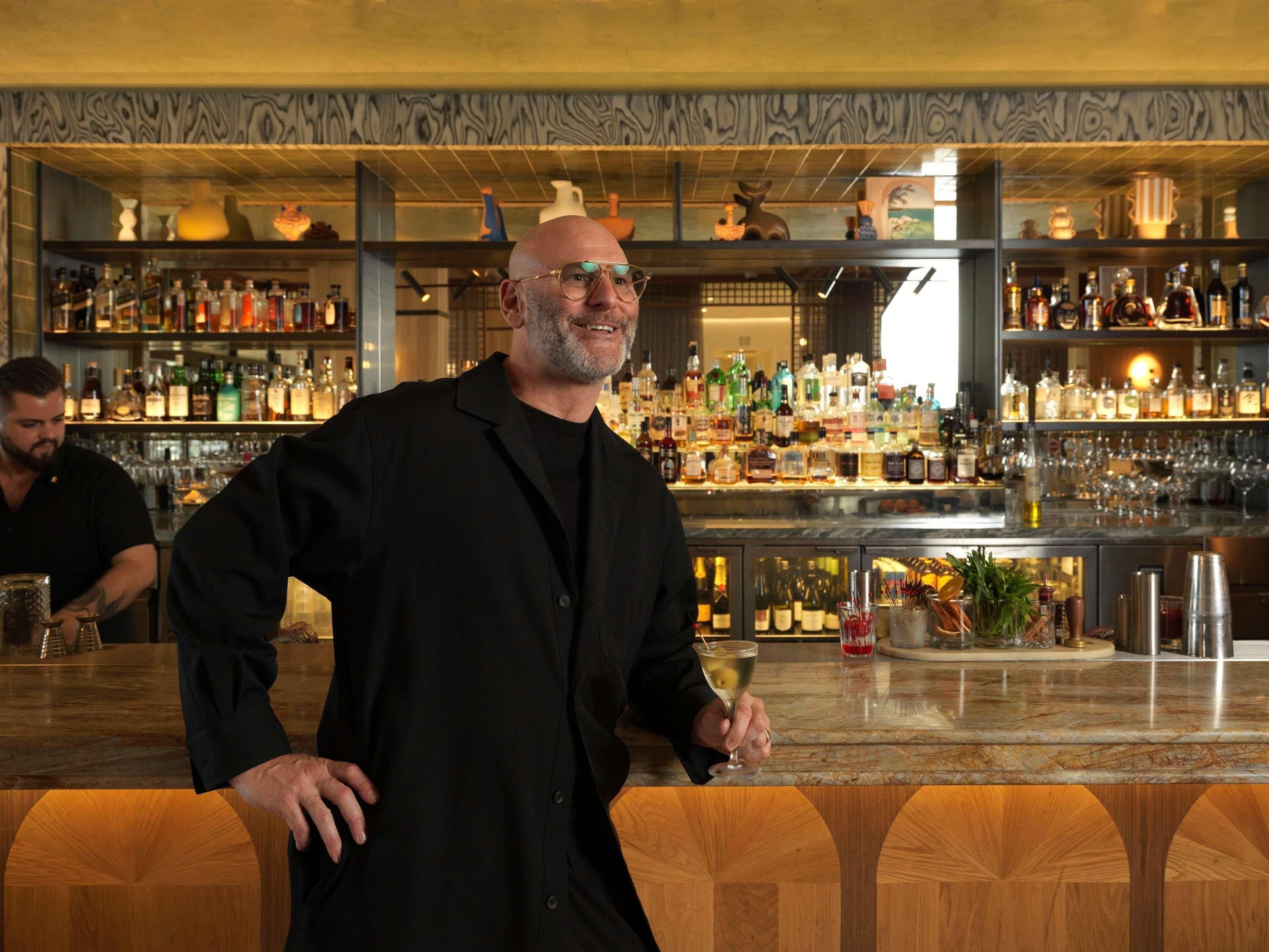 A bar area with shelves of various bottles and glasses, a marble countertop, and a person holding a drink.