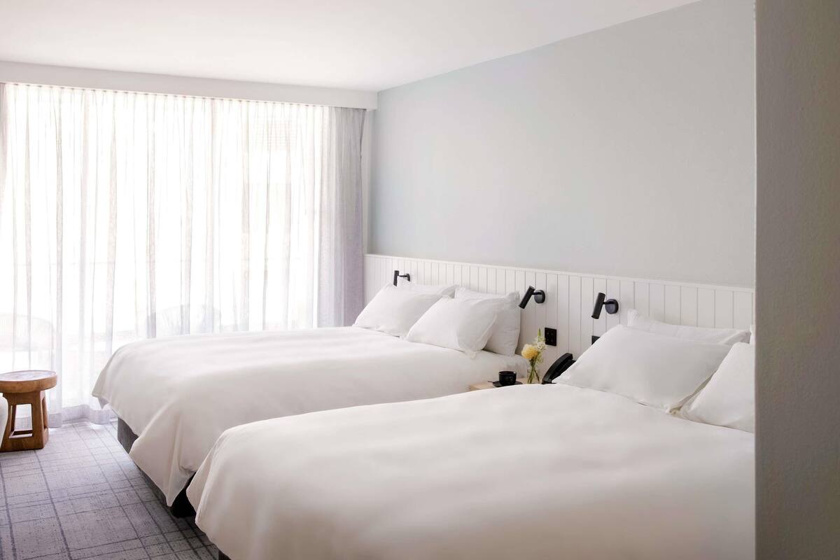 A hotel room with two beds, a small wooden stool, a bedside table with a phone and a vase of flowers, and a large window with sheer curtains.