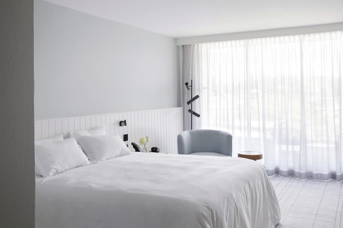 A hotel room with a large bed, a chair, a bedside table, and a window with sheer curtains.