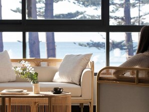 A cozy seating area with a view of the sea through large windows, a wicker chair, a cushioned sofa, and a small round table with a cup and saucer.