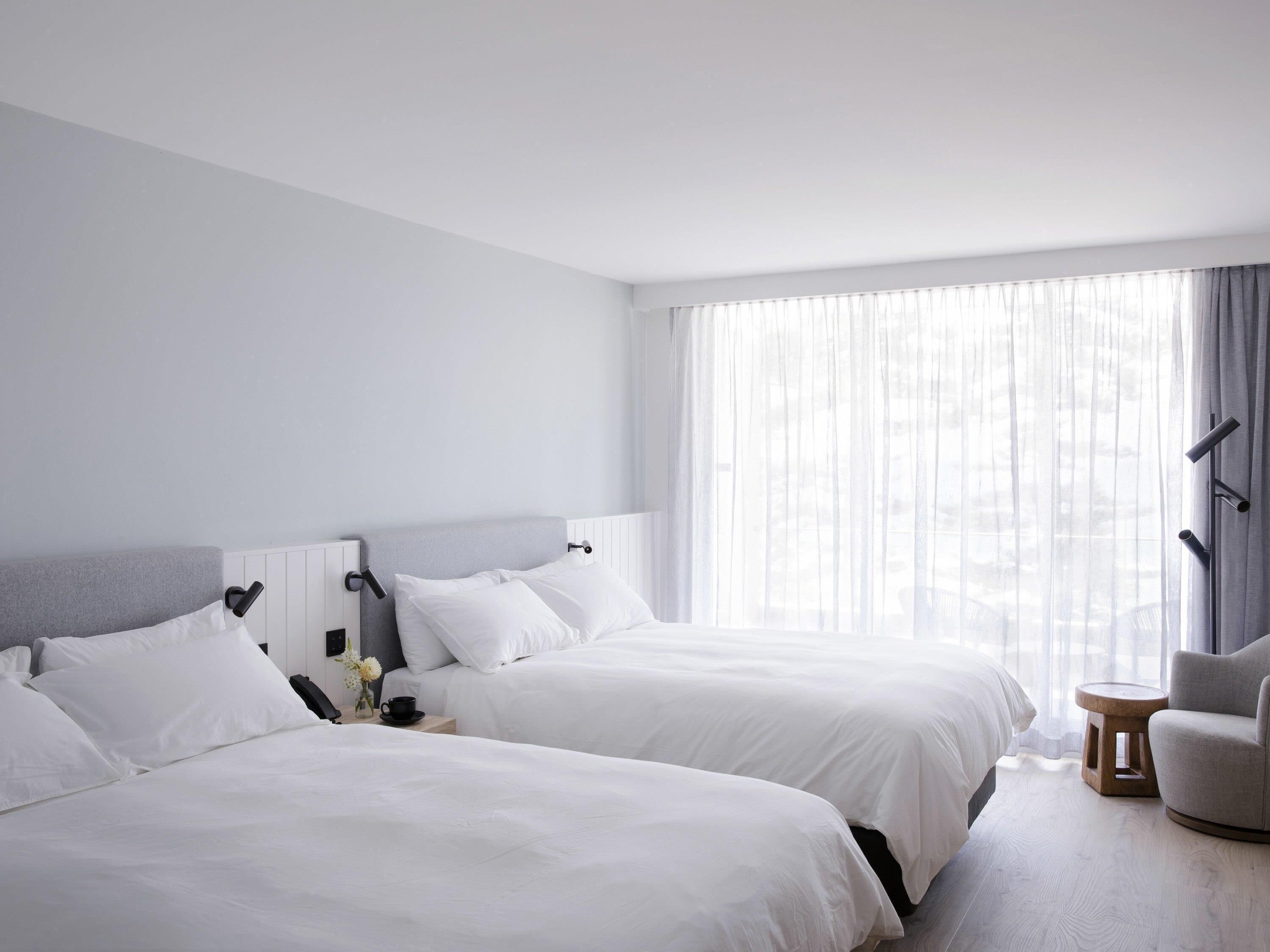 Habitación de hotel con dos camas, una mesita pequeña, una silla y un ventanal con cortinas traslúcidas.