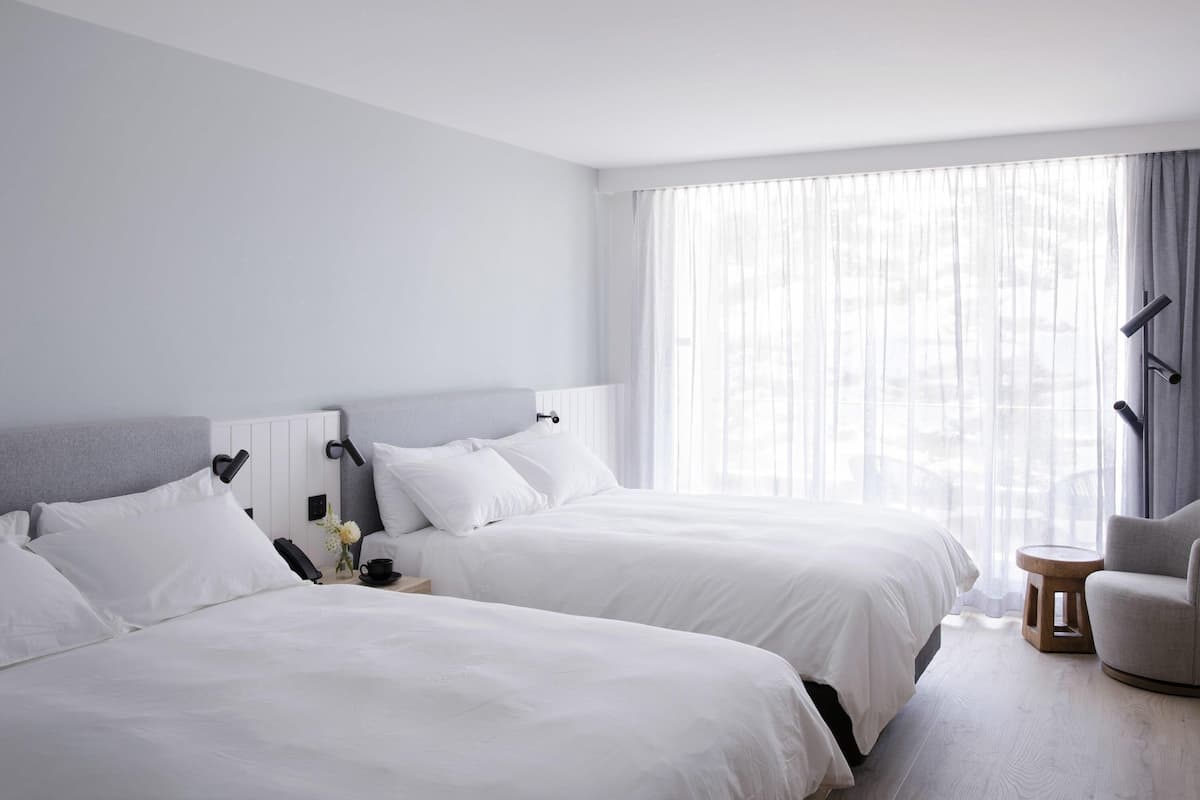 Habitación de hotel con dos camas, una mesita pequeña, una silla y un ventanal con cortinas traslúcidas.
