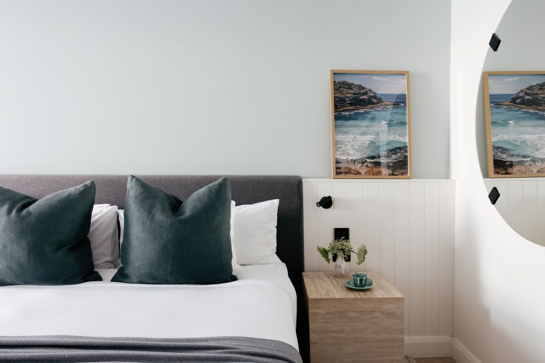 Um quarto com uma cama, travesseiros, uma mesa de cabeceira com um vaso e uma xícara, e duas quadros na parede.
