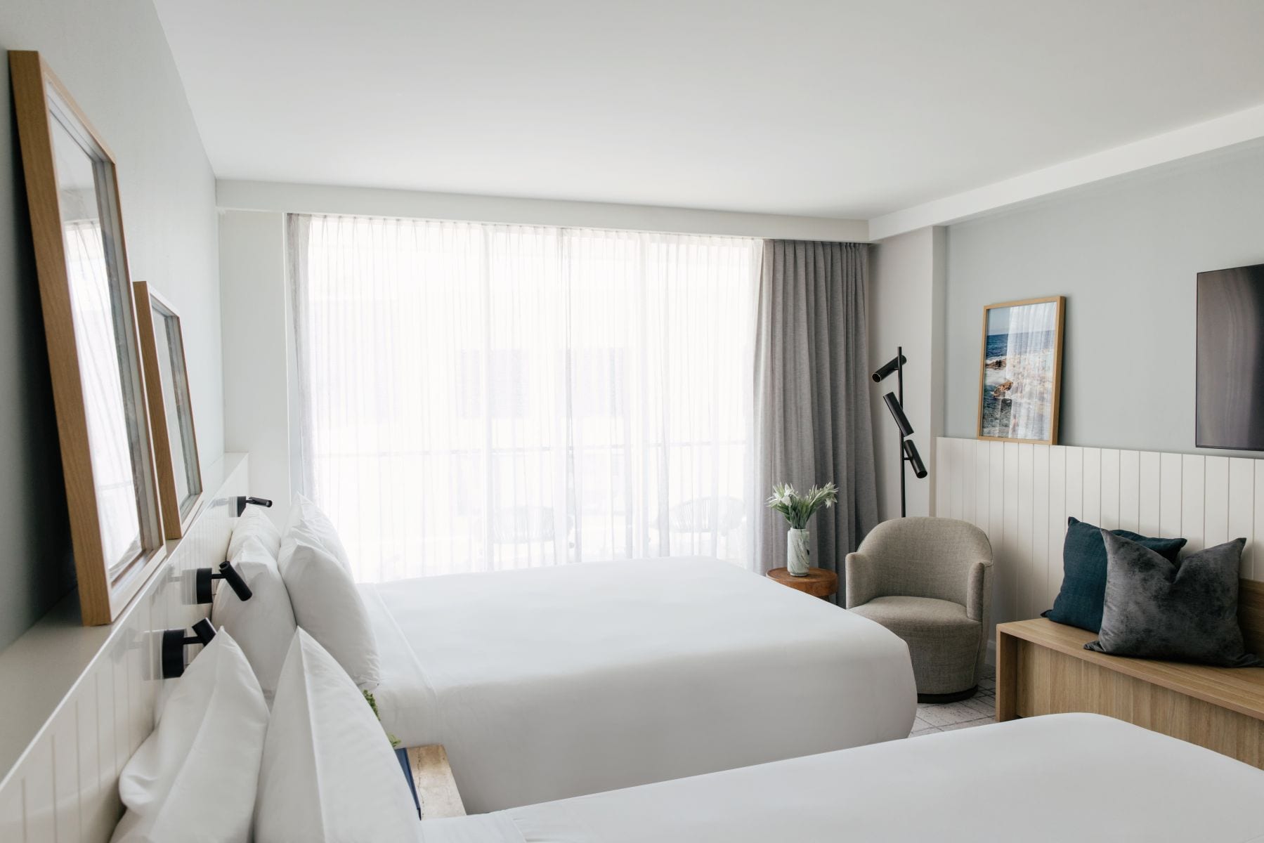 A hotel room with two beds, a chair, a nightstand, a lamp, and a window with curtains.