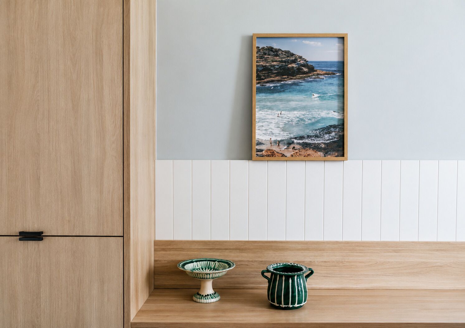 A wooden cabinet, a framed coastal landscape painting, and two ceramic containers on a wooden shelf.