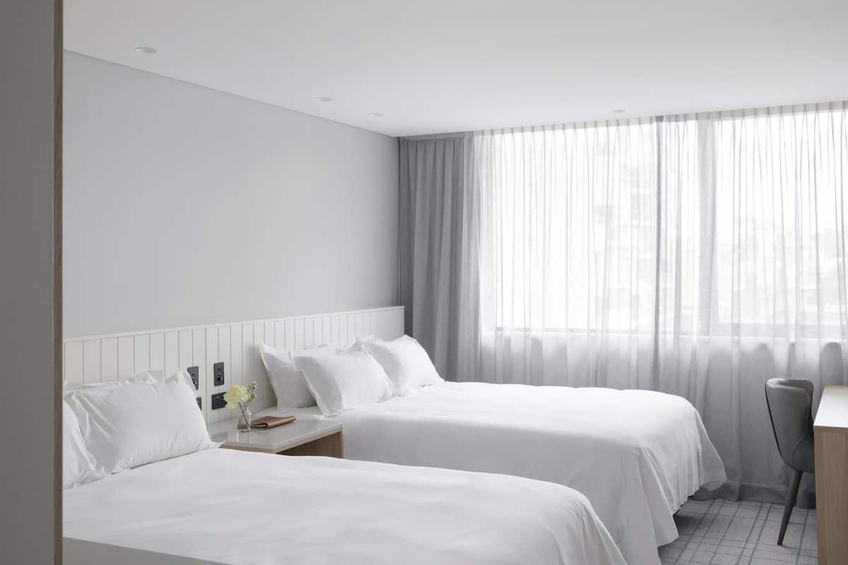 A hotel room with two beds, a desk, and a window with sheer curtains.