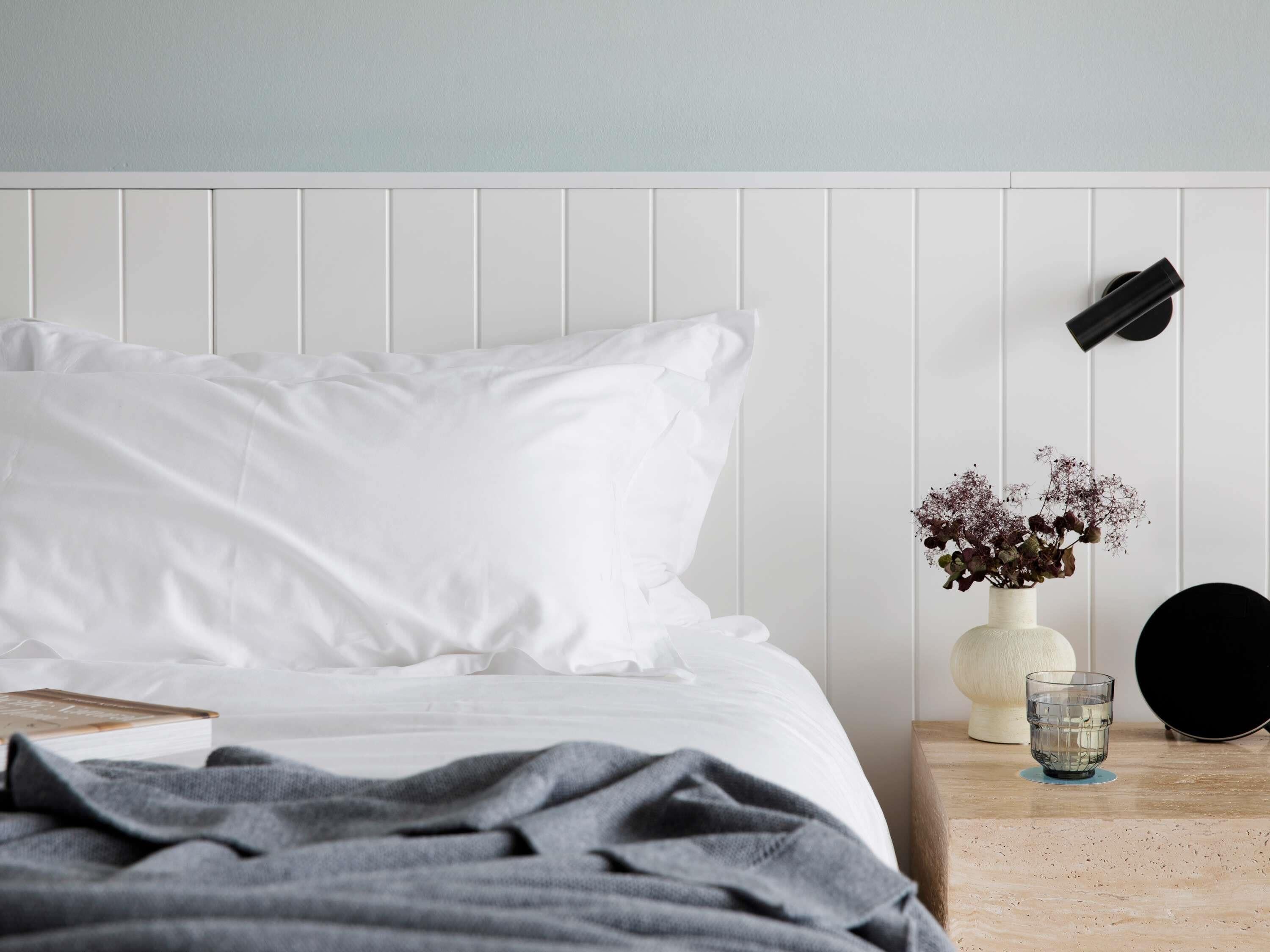 Un lit bien fait, avec une literie blanche, un plaid gris, une table de chevet ornée d’un vase de fleurs, un verre d’eau et un objet noir accroché au mur.