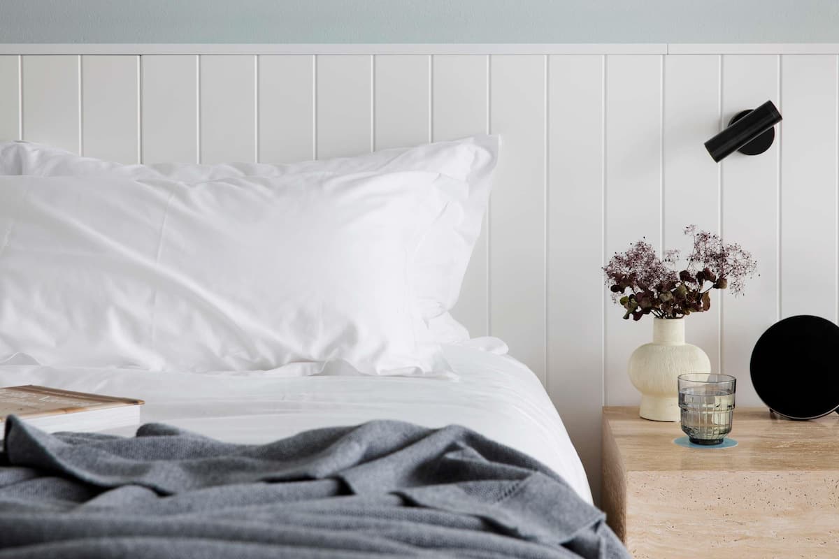Una cama bien hecha con ropa blanca, un plaid gris, una mesita de noche con un jarrón de flores, un vaso de agua y un objeto negro en la pared.