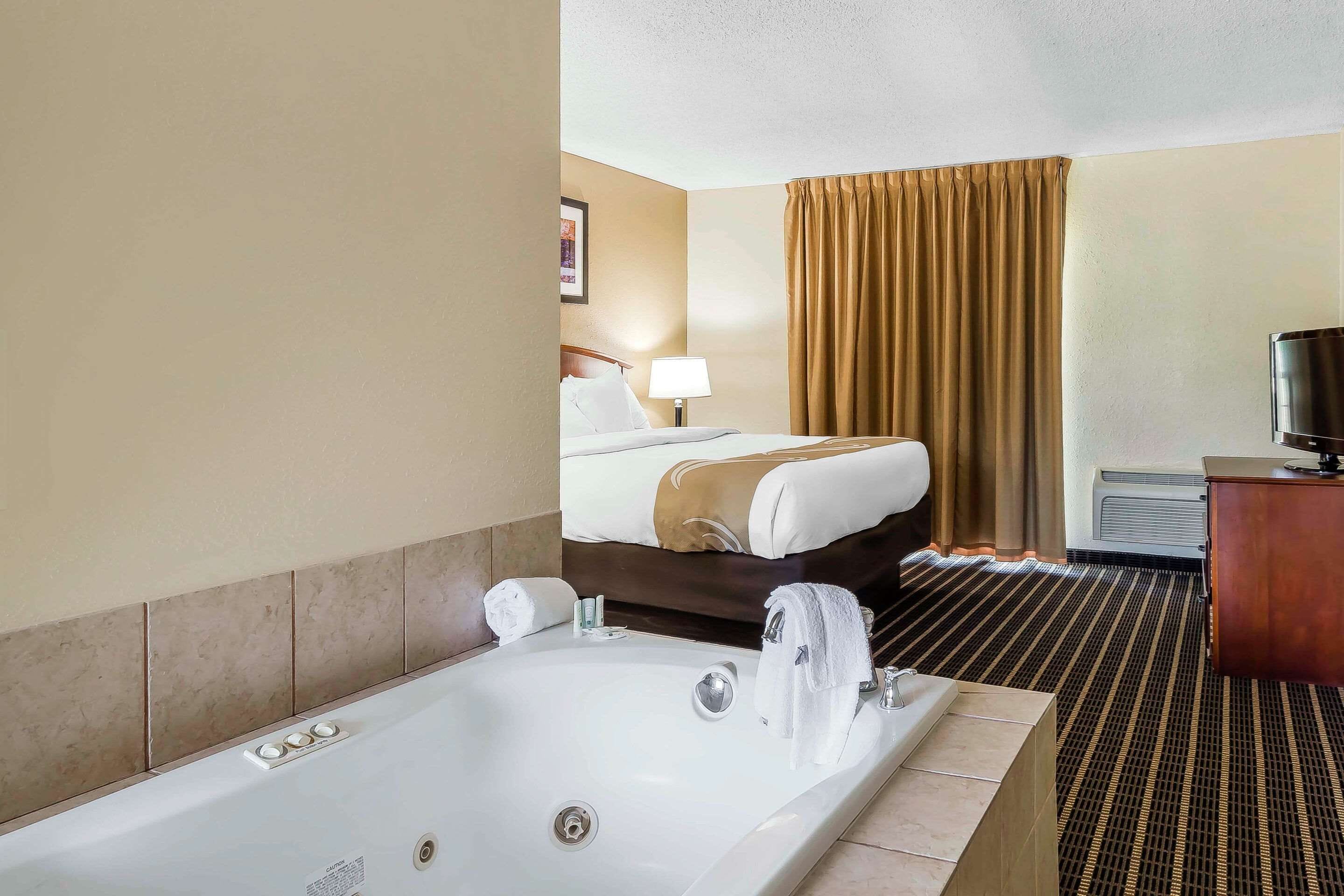 A hotel room with a large bathtub, a bed with a headboard, a television, and a desk with a chair.