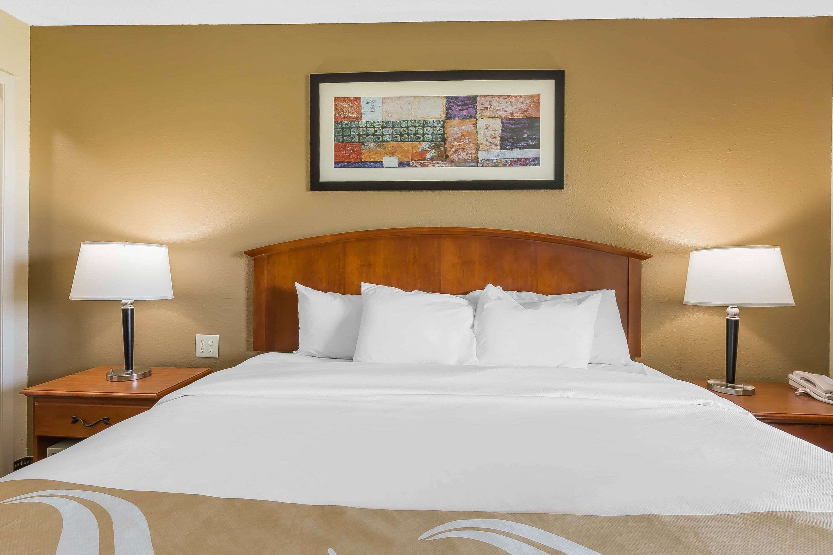 Una habitación de hotel con una cama grande, dos mesitas de noche con lámparas, un cabecero de madera y una obra enmarcada sobre la cama.