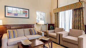 Une chambre d’hôtel comprenant un canapé, deux fauteuils, une petite table, une lampe, une fenêtre avec des rideaux et une œuvre d’art encadrée accrochée au mur.