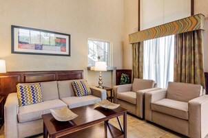 A hotel room with a sofa, two armchairs, a small table, a lamp, a window with curtains, and a framed artwork on the wall.