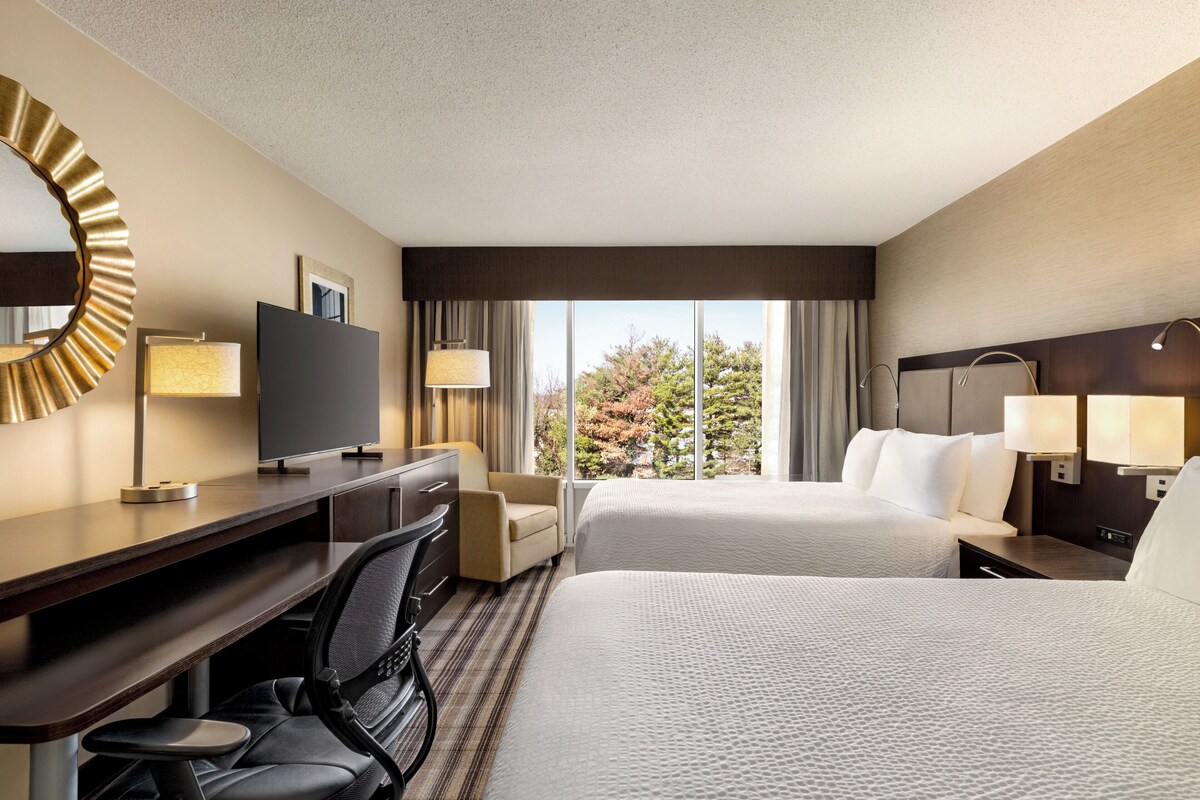 A hotel room with two beds, a desk with a chair, a TV, and a large window with a view of trees.