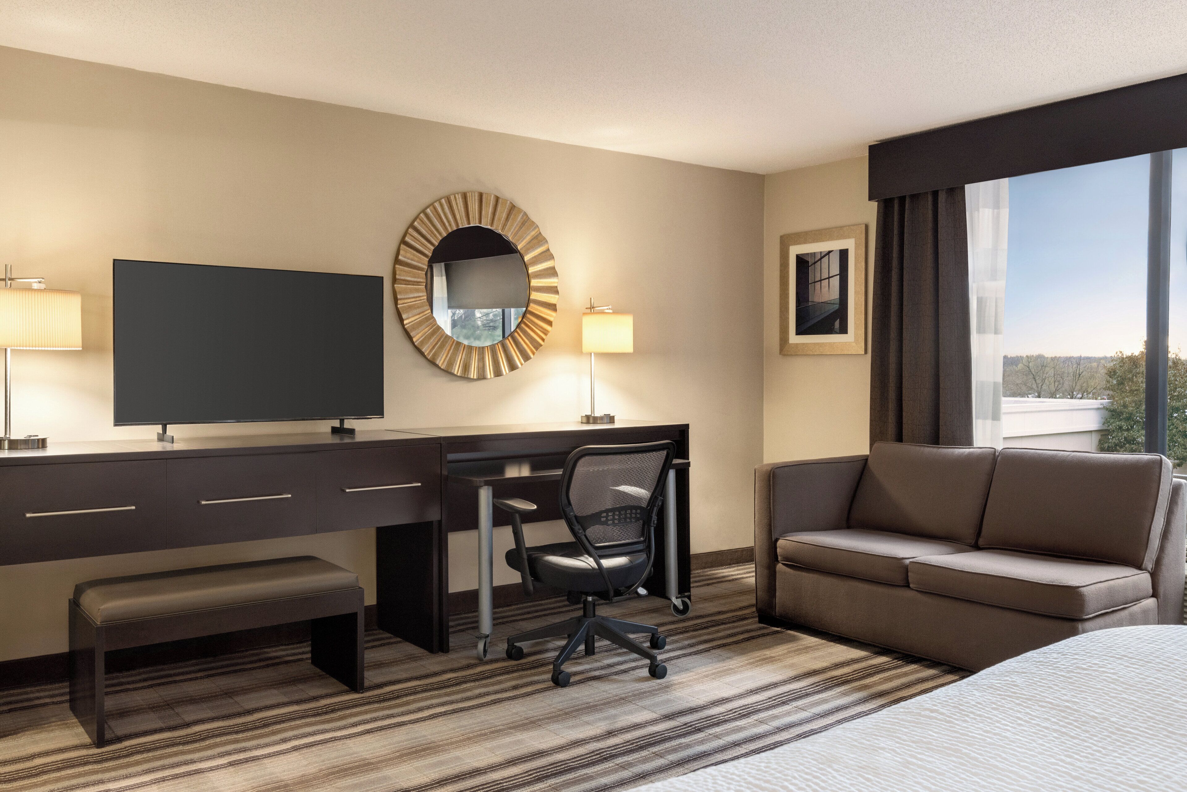 A hotel room with a large bed, a desk with a chair, a television, a round mirror, and a window with a view of the outdoors.