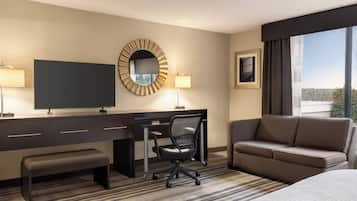 A hotel room with a large bed, a desk with a chair, a television, a round mirror, and a window with a view of the outdoors.