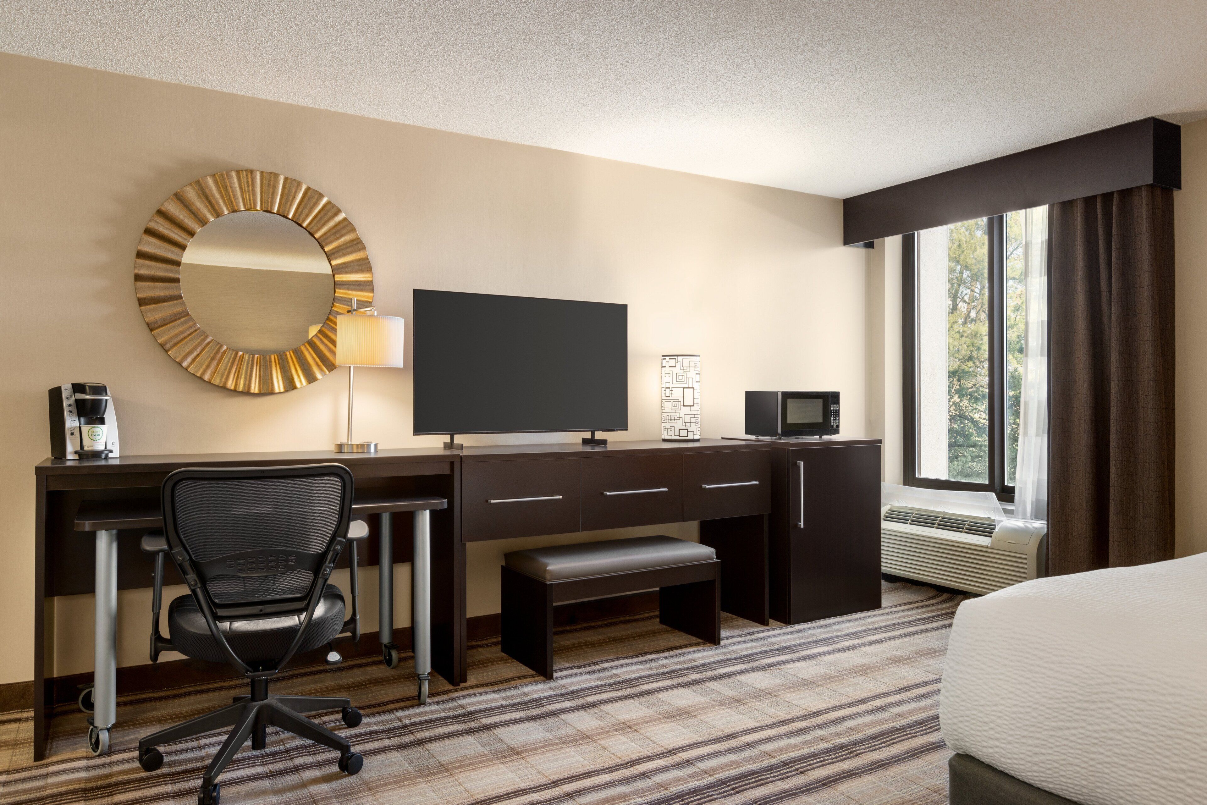 A hotel room with a bed, a desk with a computer, a chair, a large mirror, a flat-screen TV, a coffee maker, and a window with curtains.