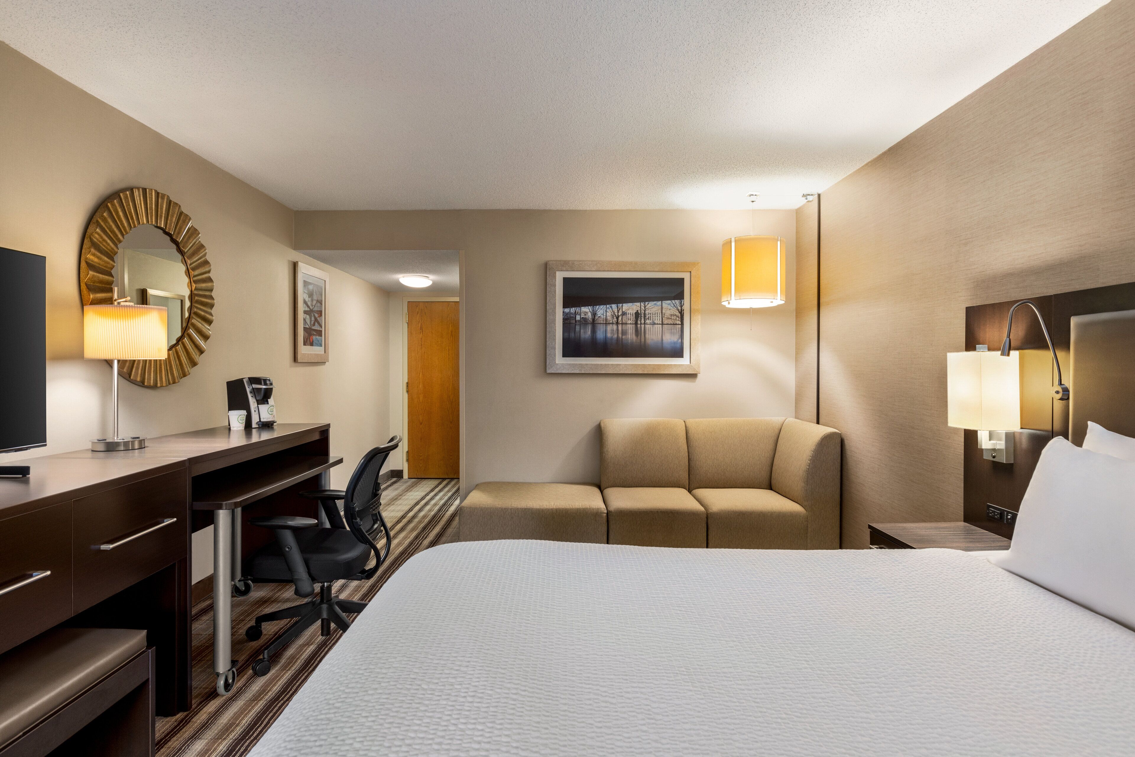 Habitación de hotel con cama, escritorio con silla, sofá, televisión y un cuadro en la pared.