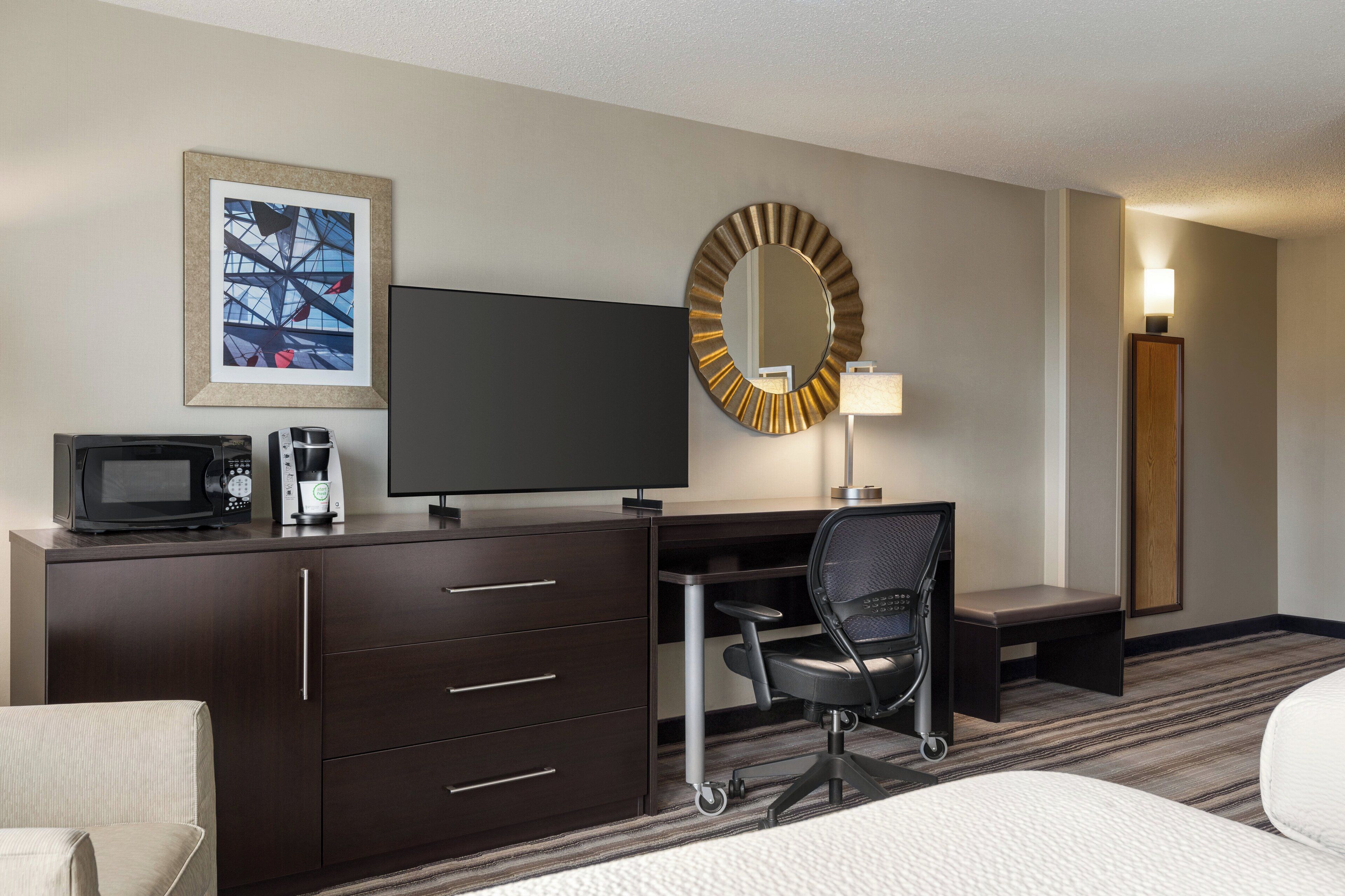 A hotel room with a bed, a desk with a chair, a television, a microwave, a coffee maker, a lamp, and a framed picture on the wall.