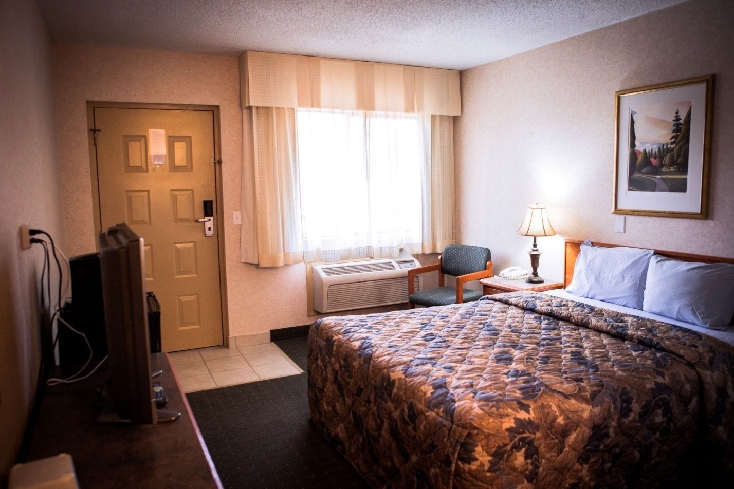 A hotel room with a bed, a desk with a lamp, a chair, a television, and an air conditioning unit.