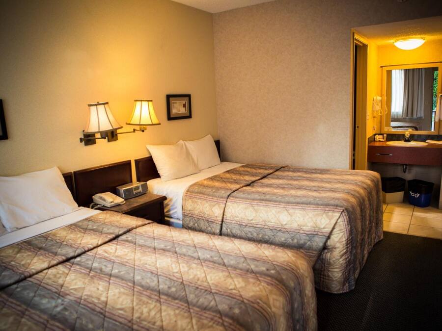 Hotellihuone, jossa on kaksi sänkyä, yöpöytä, jossa on puhelin, seinään kiinnitetty valaisin ja kylpyhuone, joka näkyy avoimesta ovesta.