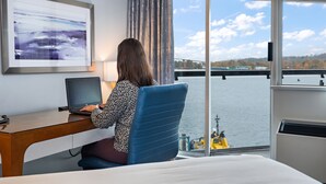 Una mujer trabajando en un escritorio con una computadora portátil, un cuadro abstracto enmarcado y vista a un río con un puente.