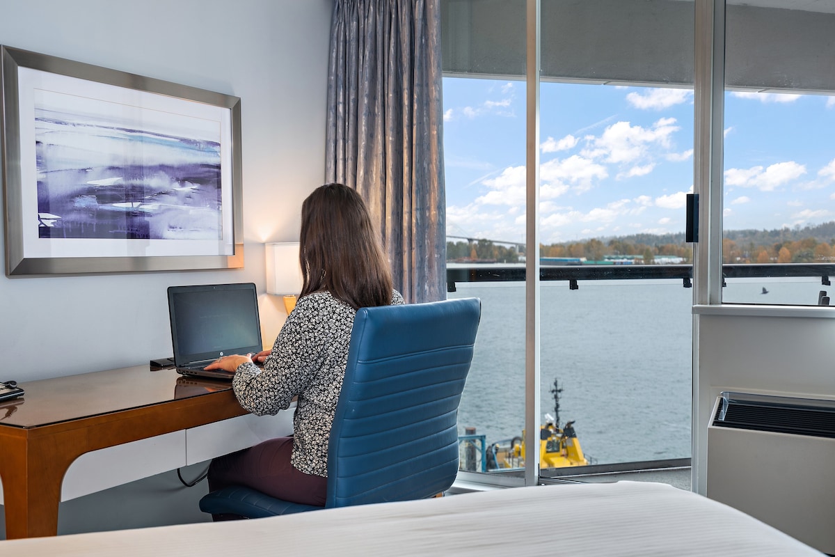 Una mujer trabajando en un escritorio con una computadora portátil, un cuadro abstracto enmarcado y vista a un río con un puente.