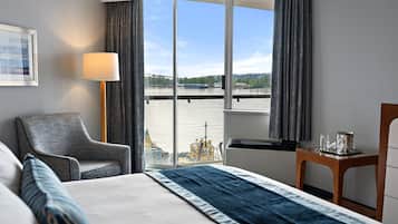 A hotel room with a bed, a chair, a table with glasses, and a view of a body of water and boats.