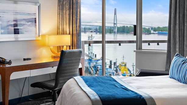 Habitación de hotel con cama, escritorio, silla, lámpara y ventana grande con vistas a un puente y un río.