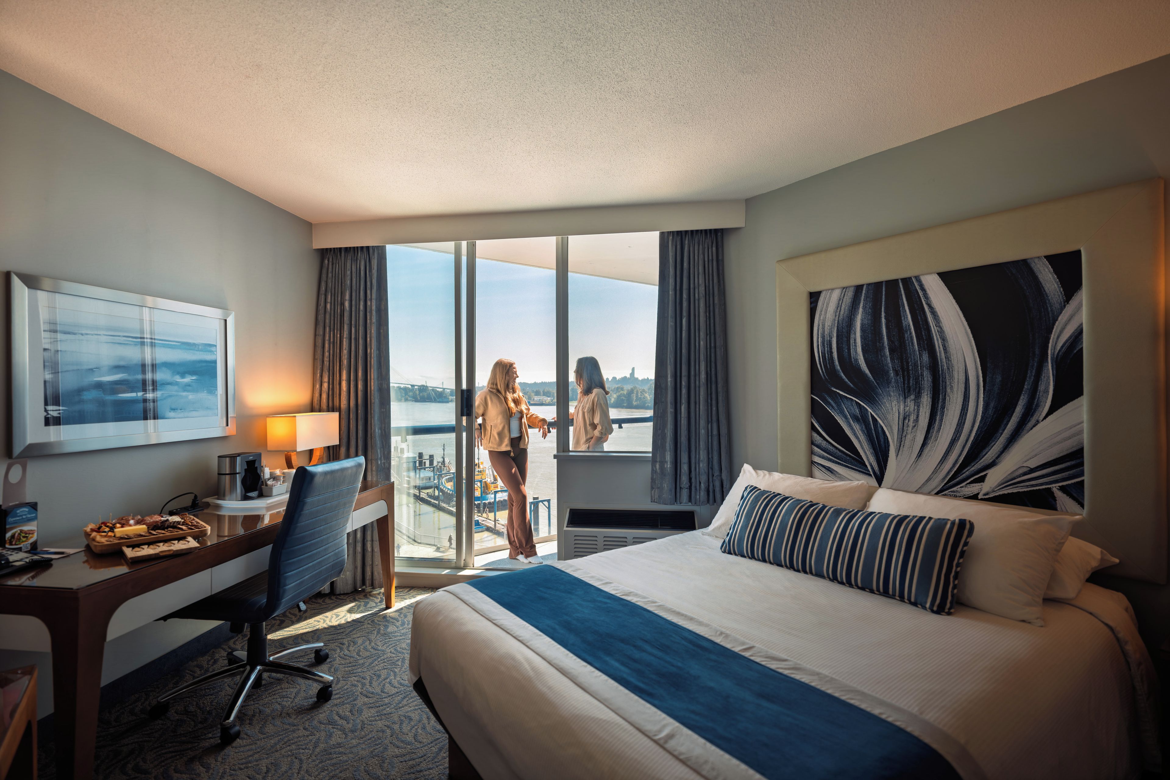 A hotel room with a large bed, a desk with a lamp, a chair, and a view of the outdoors through a sliding door.