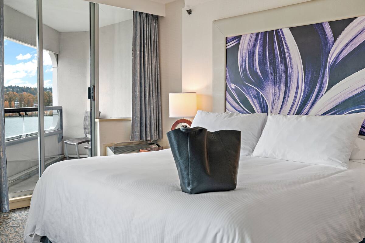 A hotel room with a large bed, a view of the outdoors, a chair, and a lamp.