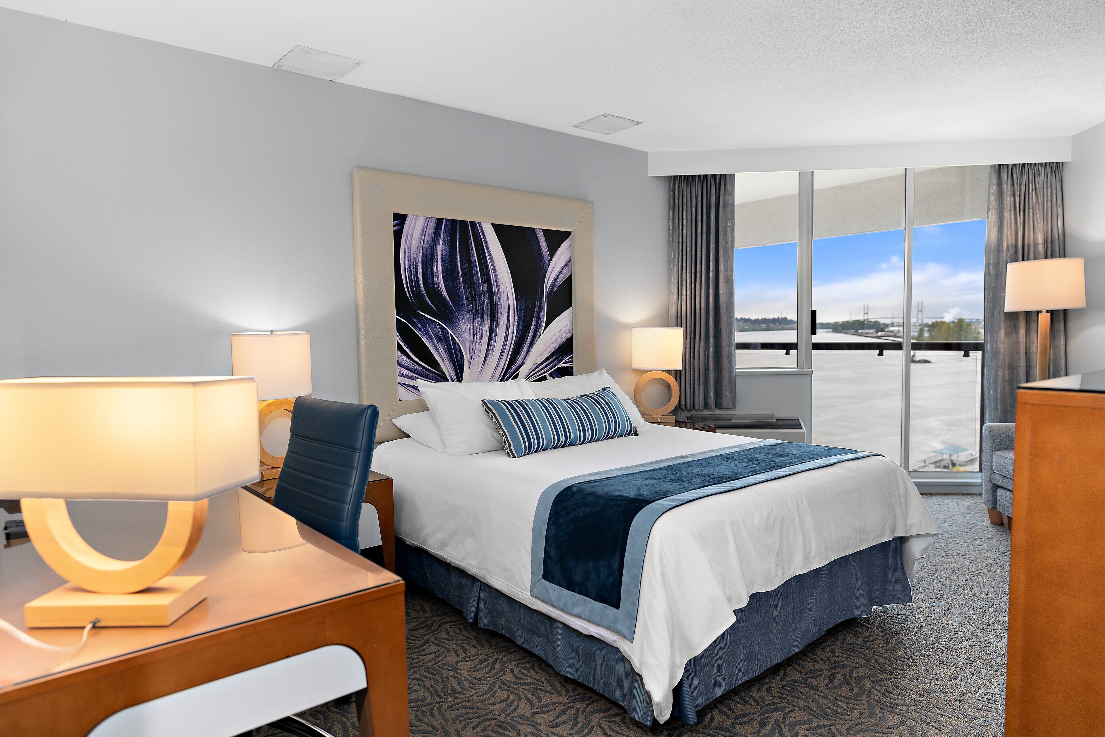 A hotel room with a large bed, two bedside tables with lamps, a desk, and a view of a bridge through large windows.