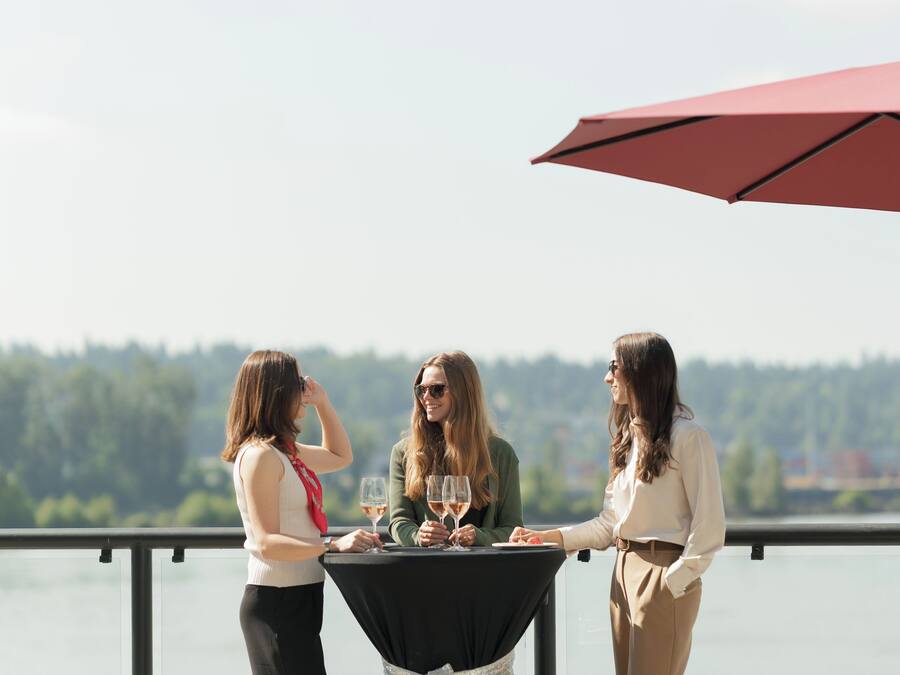 Tres personas sentadas en una mesa al aire libre, con vista a un cuerpo de agua y árboles al fondo.